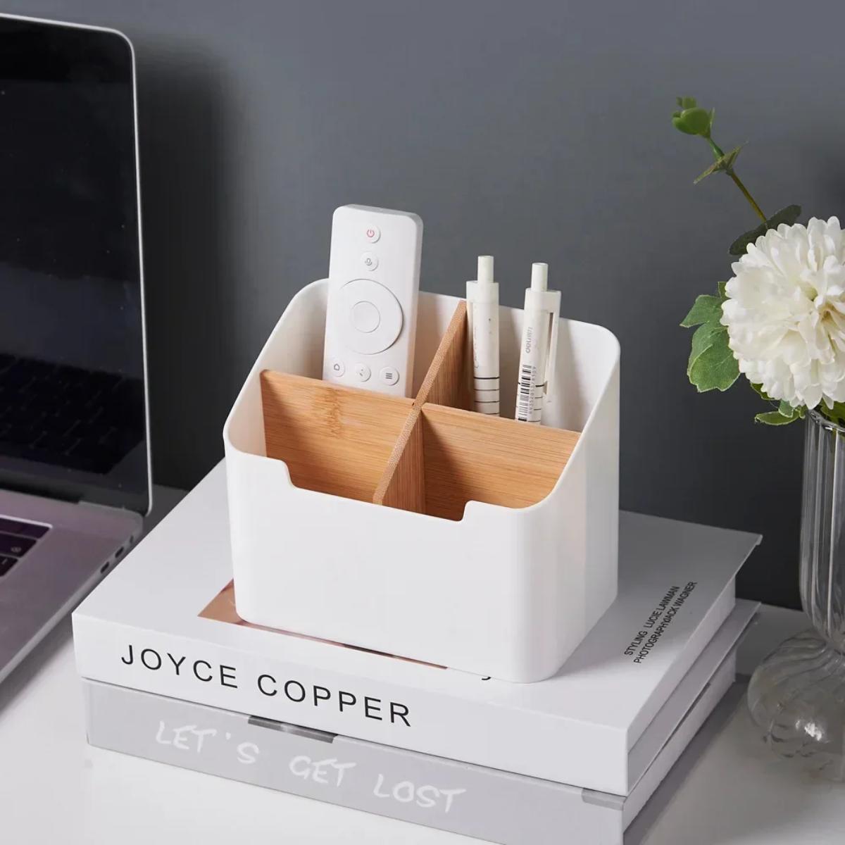 Organiseur de bureau blanc à quatre compartiments avec stylos et télécommande posés sur une pile de livres.